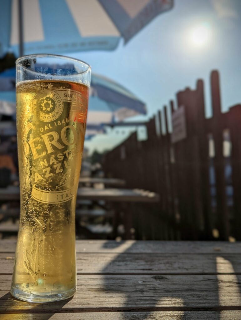 A tall glass of Peroni beer on a wooden picnic table in a sunny beer garden under blue and white parasols.