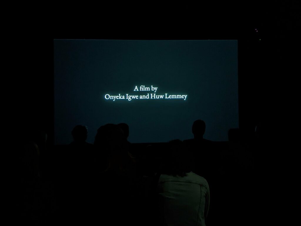 Audience silhouettes in a dark screening room watching a title card that reads: A film by Onyeka Igwe and Huw Lemmey