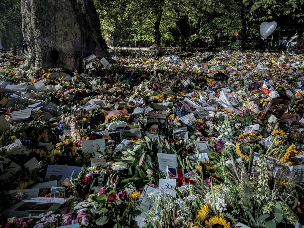 A vast sea of floral tributes, cards, and portraits of Queen Elizabeth II surrounding a tree in a park at night.