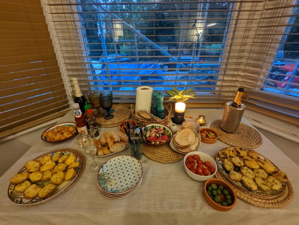 A variety of appetizers including bruschetta, olives, and salad spread across a table with champagne by a window at dusk.