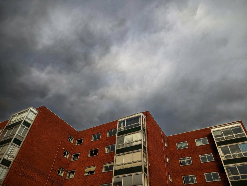 A red brick apartment block with glass balconies under a heavy, swirling dark grey storm sky.