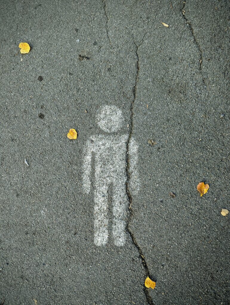 White stenciled human figure on grey asphalt, split by a vertical crack and surrounded by fallen yellow leaves.