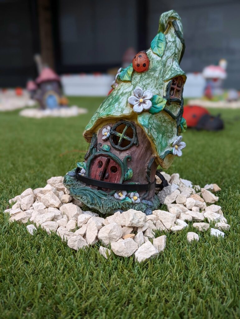 A whimsical fairy house garden ornament with a leafy green roof and red door, nestled on white gravel in a lush green lawn.