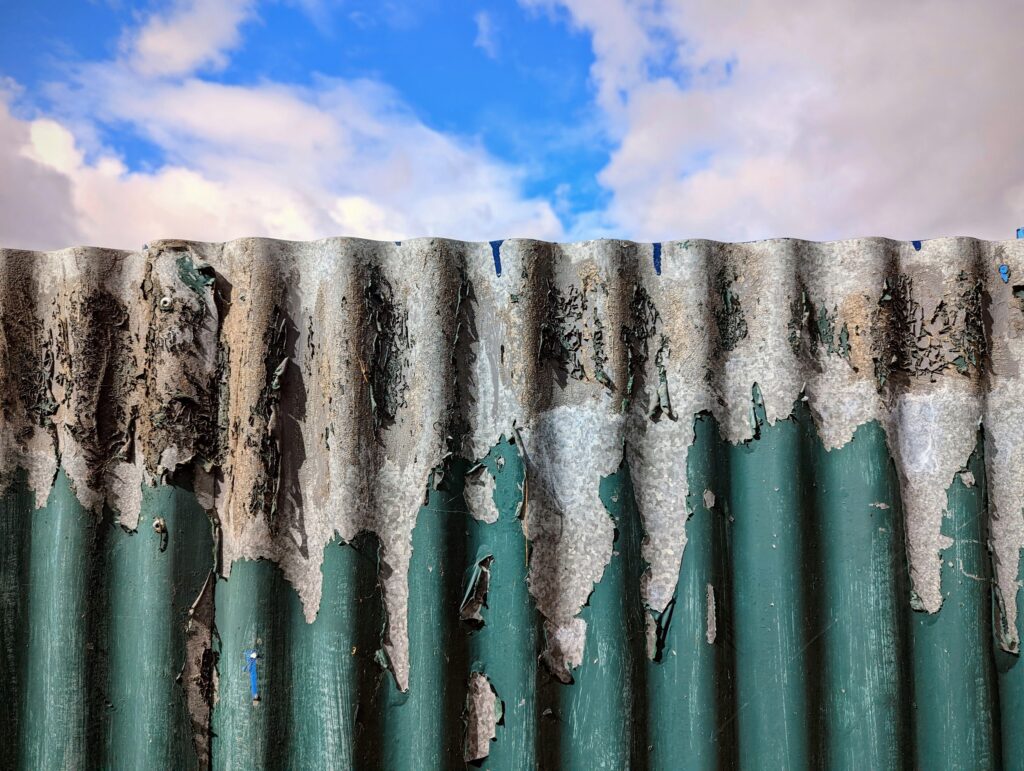 Weathered corrugated metal fence with peeling green paint against a bright blue sky with fluffy white clouds.