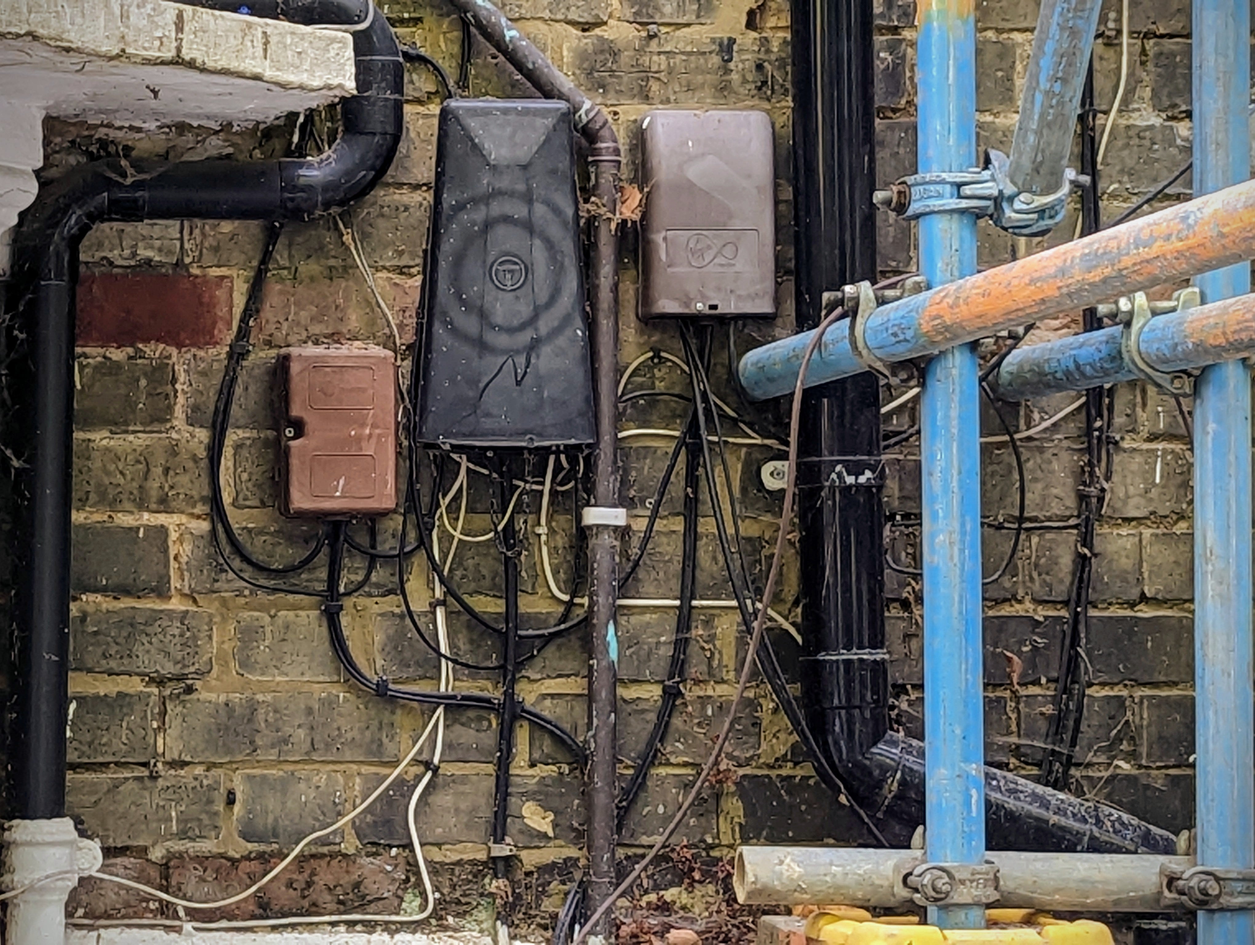 Virgin Media and Openreach junction boxes with tangled wiring on a brick wall next to black pipes and blue scaffolding poles.