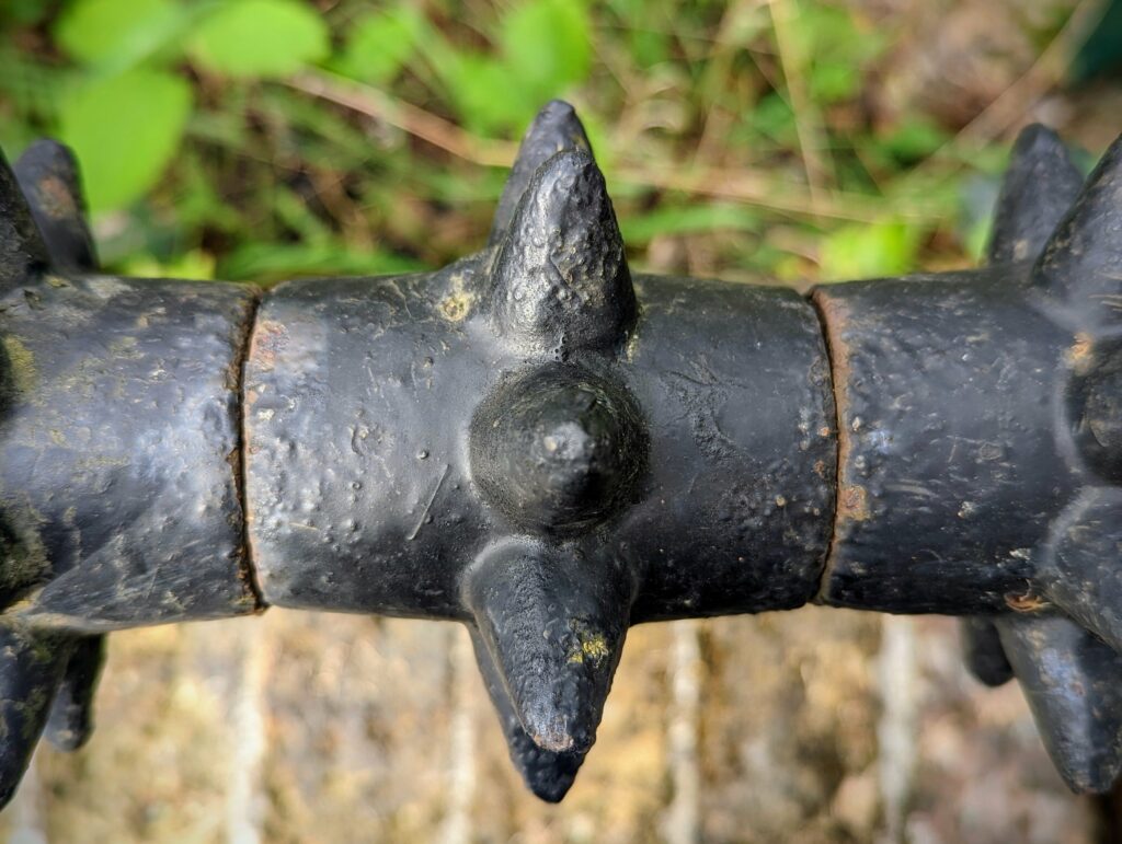 A weathered black iron railing with thick, rounded spikes against a blurred background of green foliage and a stone wall.