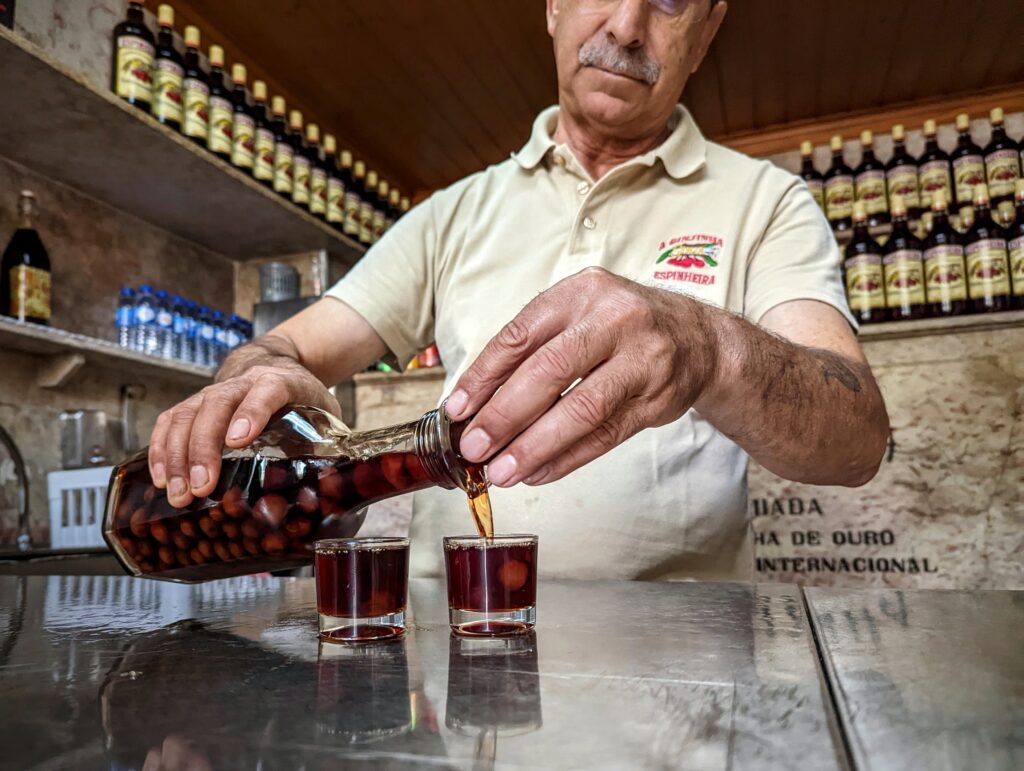 A bartender at A Ginjinha Espinheira pours cherry liqueur from a bottle containing whole fruit into two shot glasses.