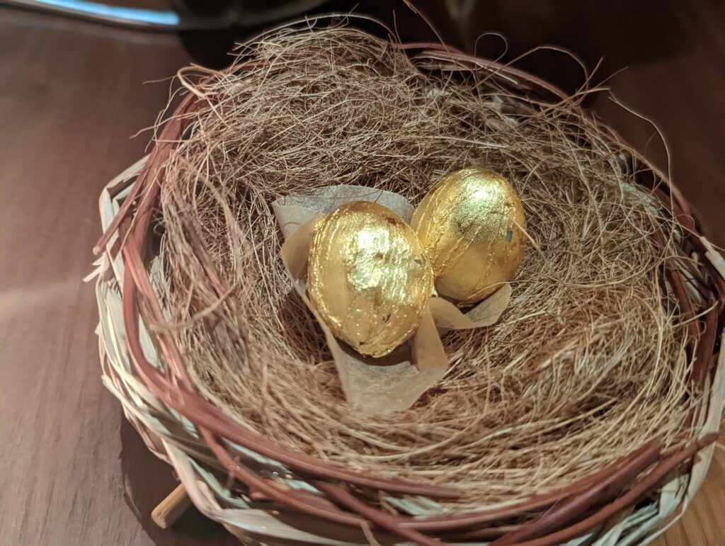 Two gold-wrapped eggs nestled inside a small decorative bird's nest made of twigs and straw.