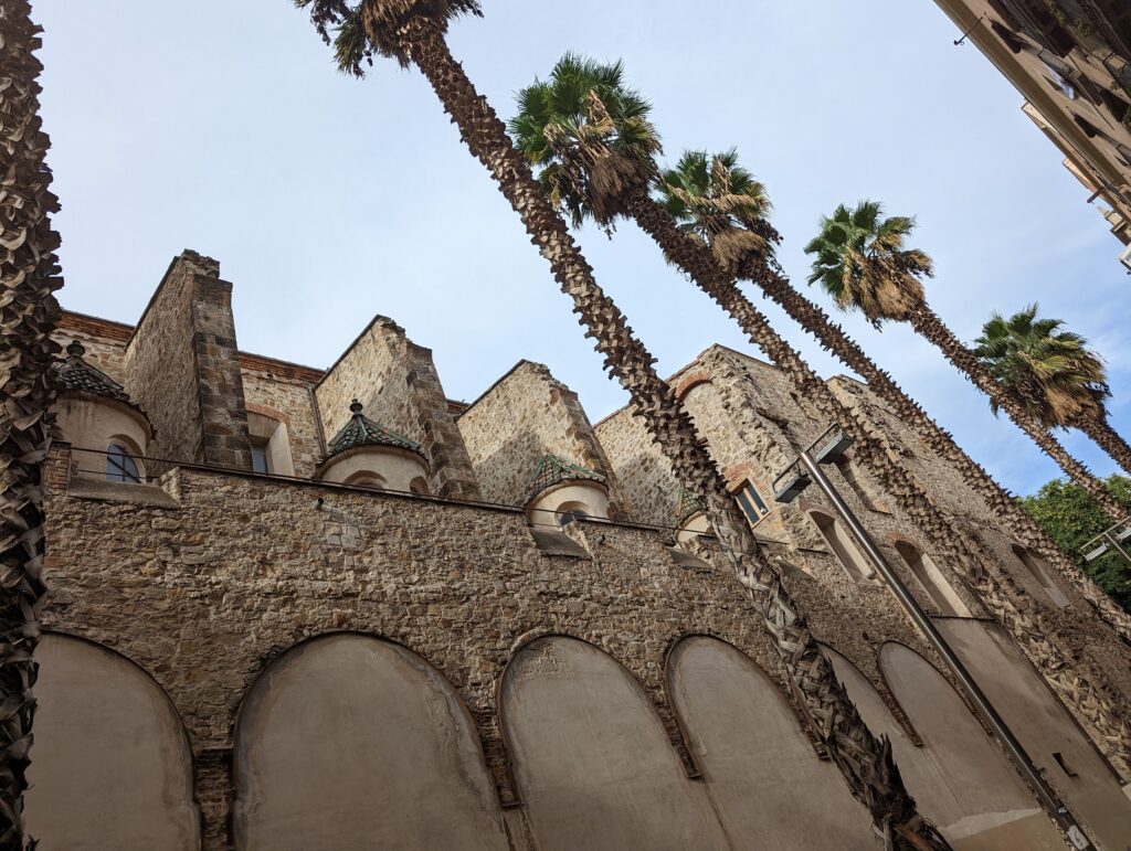 Tall palm trees leaning over an ancient stone wall with arched alcoves and small tiled domes under a pale blue sky.