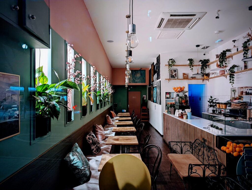Long cushioned bench seating and wooden tables inside a cozy cafe with pink and green walls, hanging bulbs, and indoor plants.