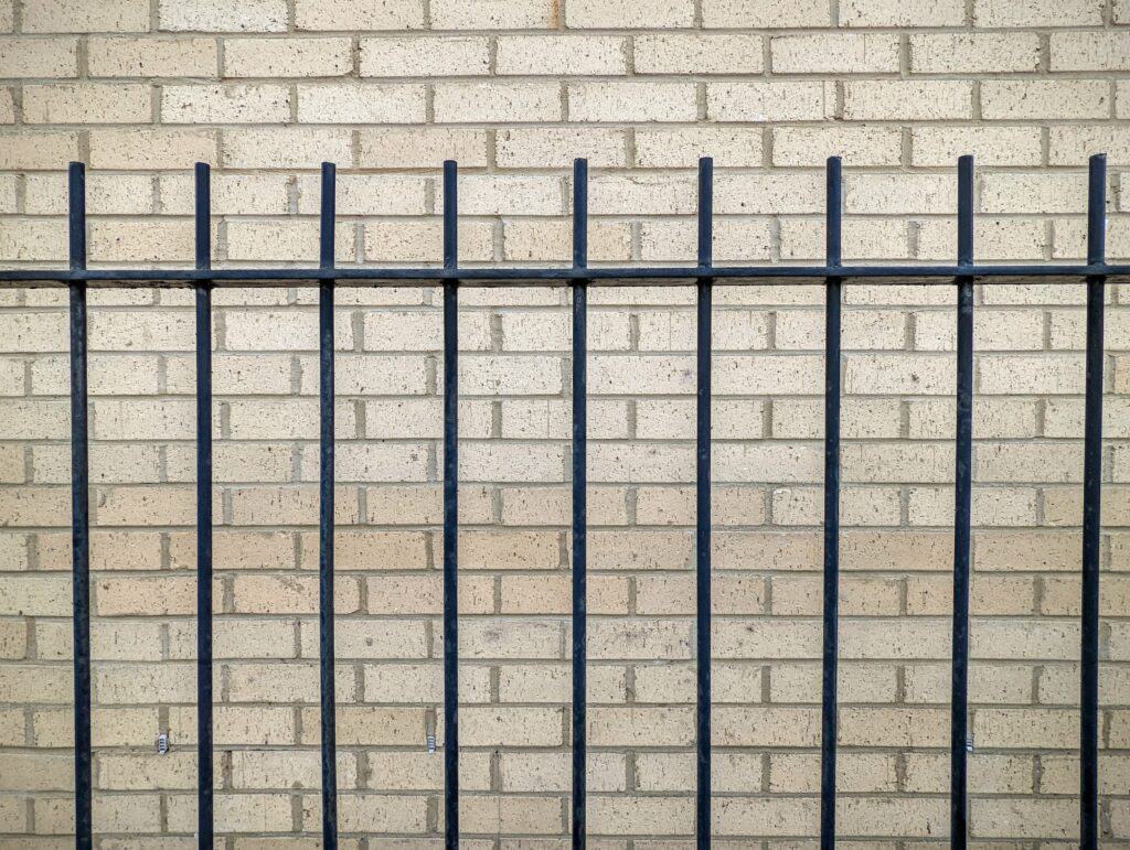 Vertical black metal railings positioned in front of a textured light beige brick wall.