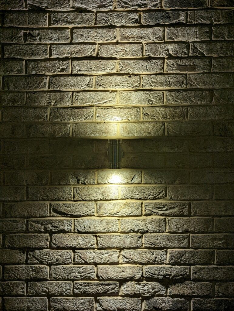 A textured brick wall illuminated by a cylindrical wall light casting warm beams of light upwards and downwards.