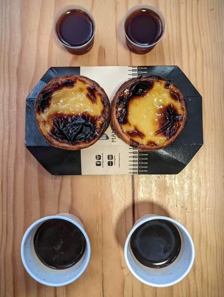 Two golden-brown pastel de nata in a branded holder, flanked by four cups of coffee on a light wooden table, captured from directly above.