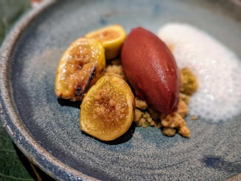 Halved roasted figs and a red sorbet quenelle atop a crumble base with white foam, served on a speckled blue ceramic plate.