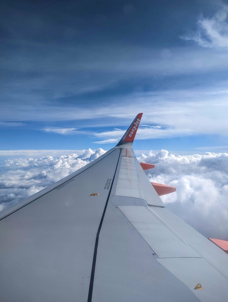 An easyJet airplane wing with an orange winglet soaring above a dense layer of white clouds under a blue sky.