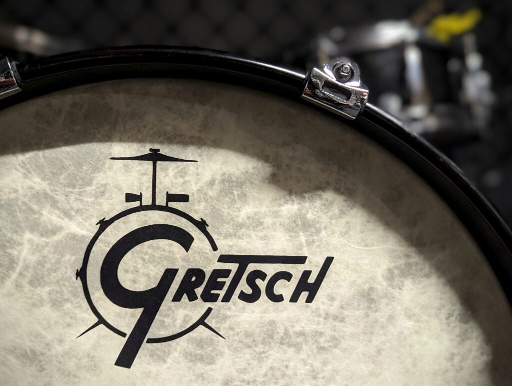 A close-up of a Gretsch drum head with a textured, parchment-like finish and black logo, featuring a dark rim and chrome lug.