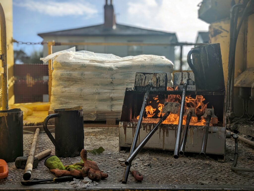 Metal rods heating in an open fire box on a truck bed, surrounded by black pouring pitchers, work gloves, and bags of road marking material.
