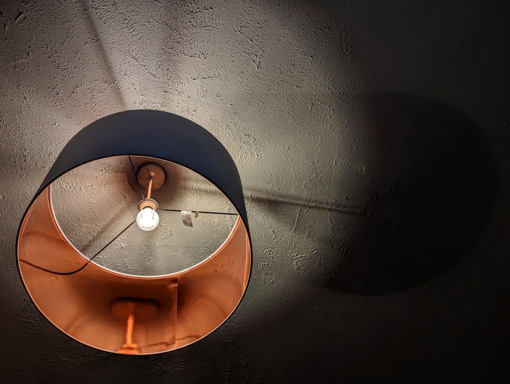 Looking up into a pendant lamp with a copper-lined interior and lit bulb, casting a large circular shadow on a textured white ceiling.