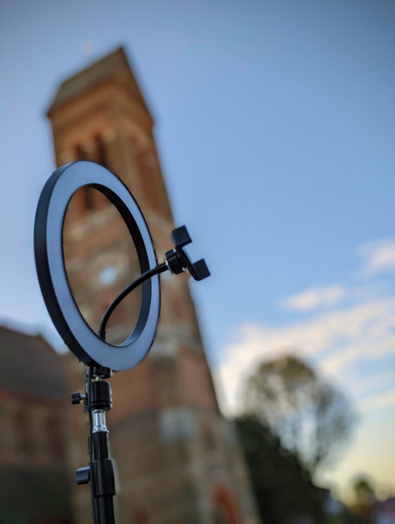A black ring light on a tripod in the foreground, set against a blurred tall brick tower and a clear blue sky.