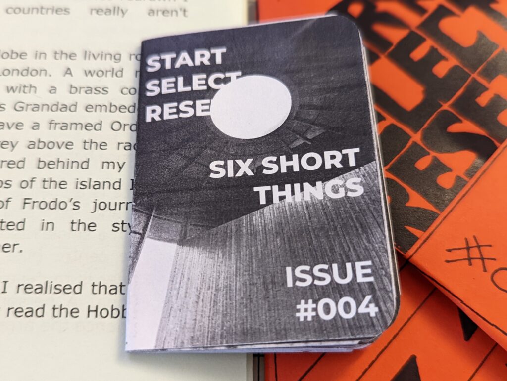 Start Select Reset zine titled Six Short Things Issue #004 with a monochrome architectural cover, resting on an open book beside bright orange pamphlets.