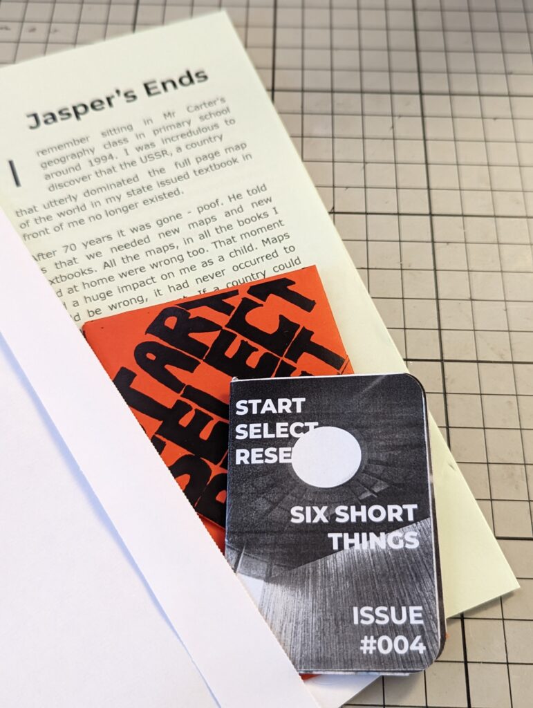 Six Short Things Issue #004 zine resting on an orange card and a page titled Jasper's Ends on a grey gridded cutting mat.