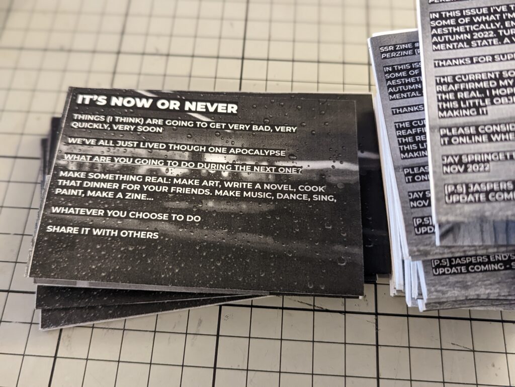Stacks of black and white zines on a gridded mat. The top card titled IT'S NOW OR NEVER urges the reader to make something real and share it.