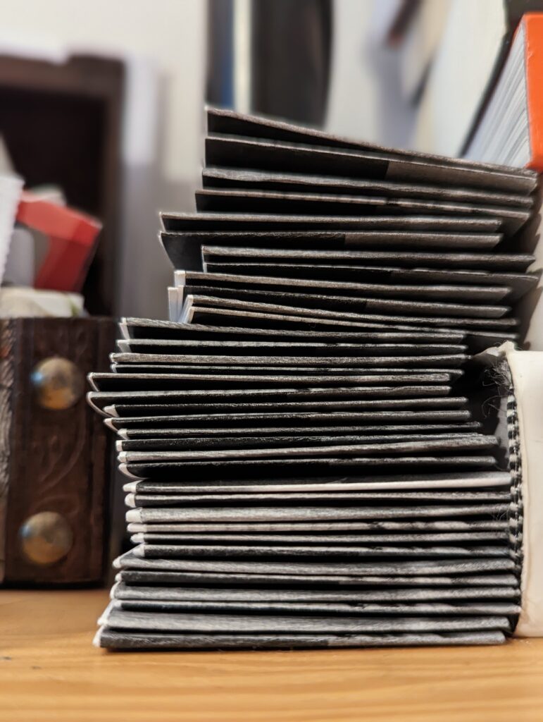 A stack of thin black zines showing white page edges on a wooden surface next to a carved dark wood box.