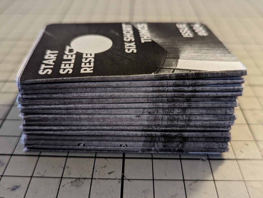 A stack of black and white zines titled Six Short Things Issue #004 sitting on a gridded cutting mat.