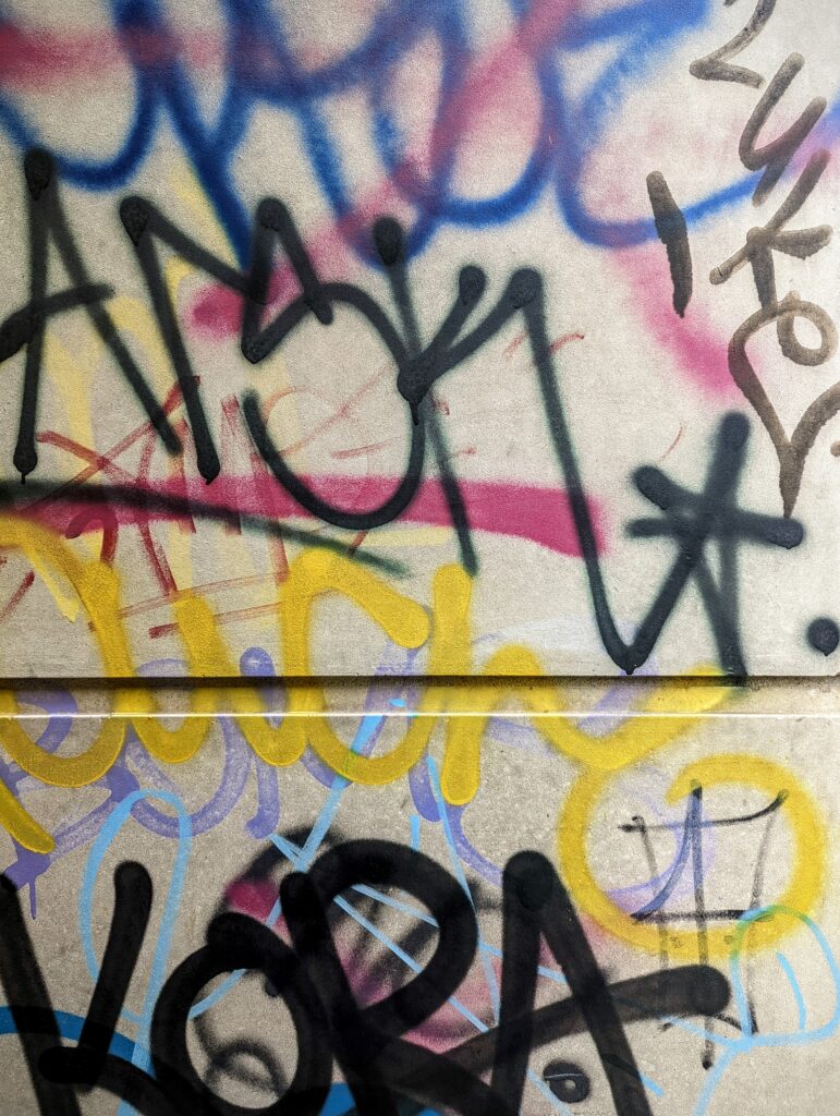 Multicoloured graffiti tags in black, yellow, pink and blue spray paint layered on a concrete wall divided by a horizontal groove.