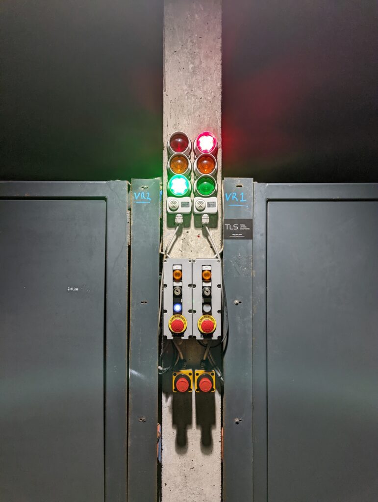 Industrial traffic lights and control panels on a concrete pillar between grey doors. One green and one red light are illuminated.