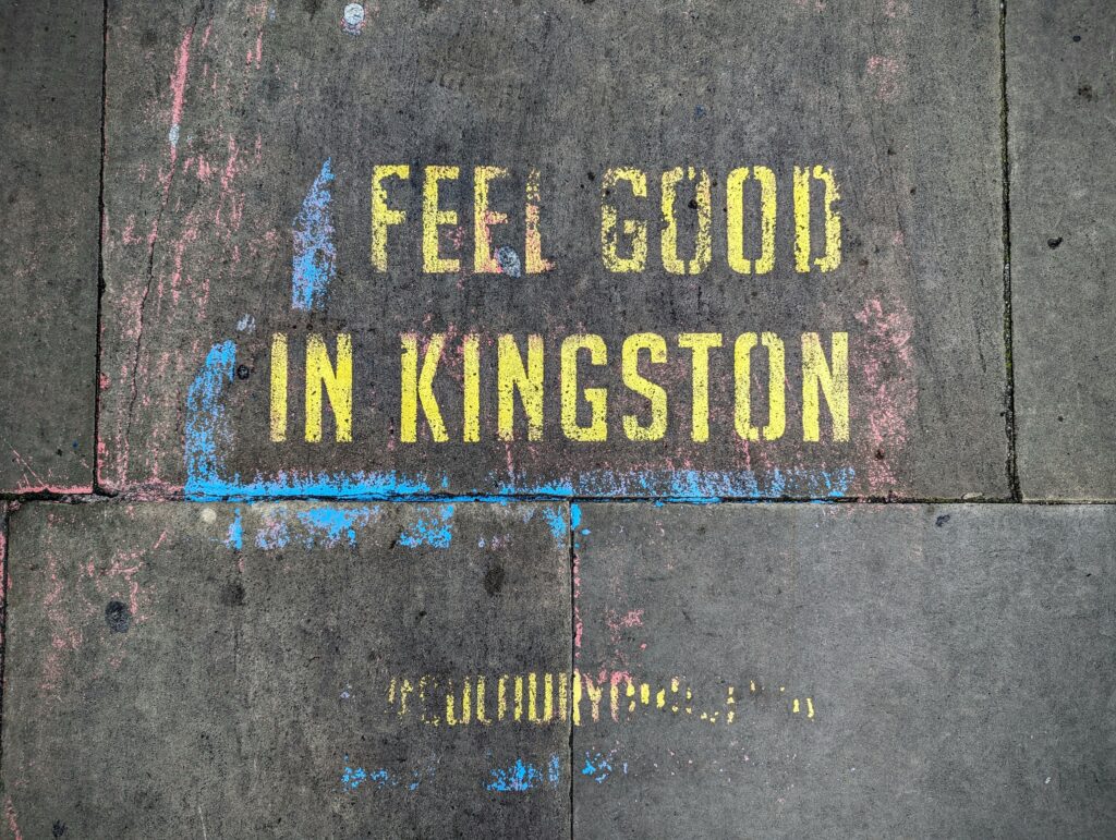 Yellow stenciled text on grey paving stones reading FEEL GOOD IN KINGSTON, surrounded by faint blue and pink chalk marks.
