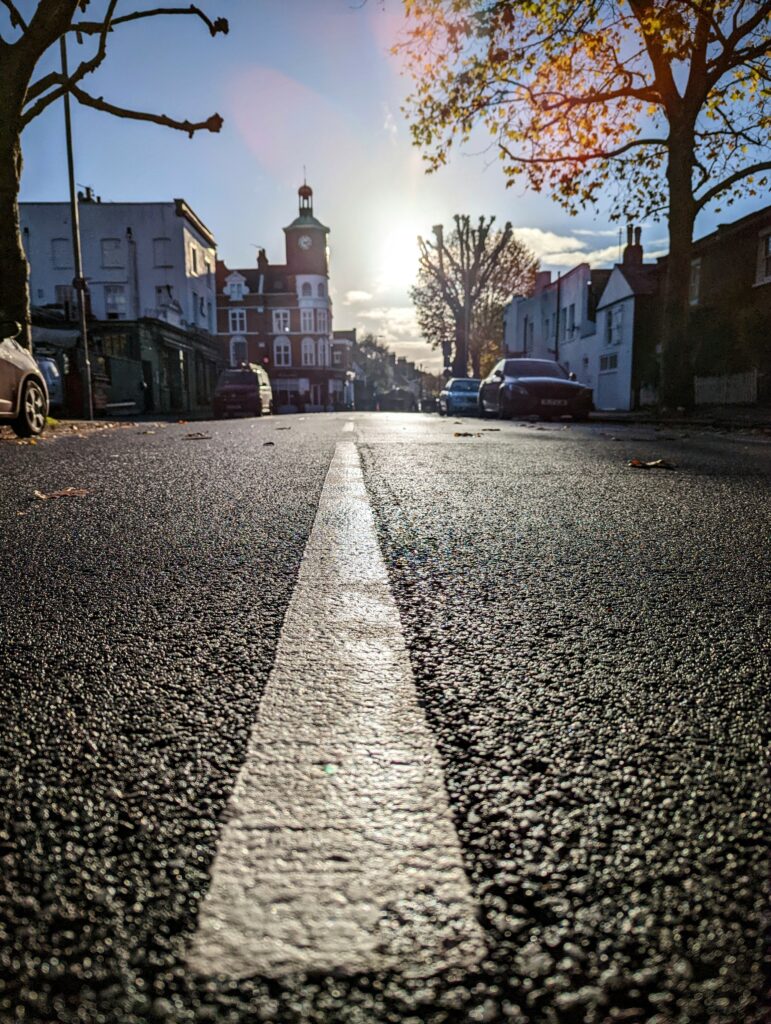 A low-angle view of a white lane marker on a paved street leading toward a brick clock tower under a bright, low autumn sun.