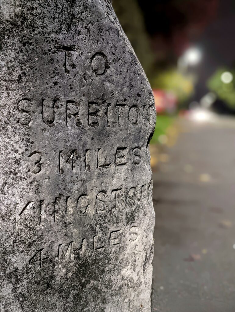 Weathered stone milestone at night engraved with TO SURBITON 3 MILES and KINGSTON 4 MILES, against a blurred street background.