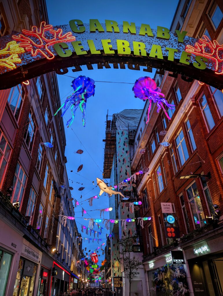 Glowing jellyfish and a shark suspended above Carnaby Street at night, beneath an illuminated arch reading "CARNABY CELEBRATES".