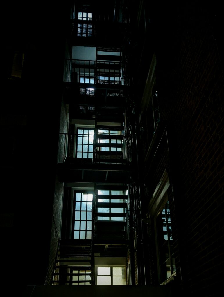 Silhouetted metal fire escape rising up a dark building at night, with glowing window panes lighting each landing level.