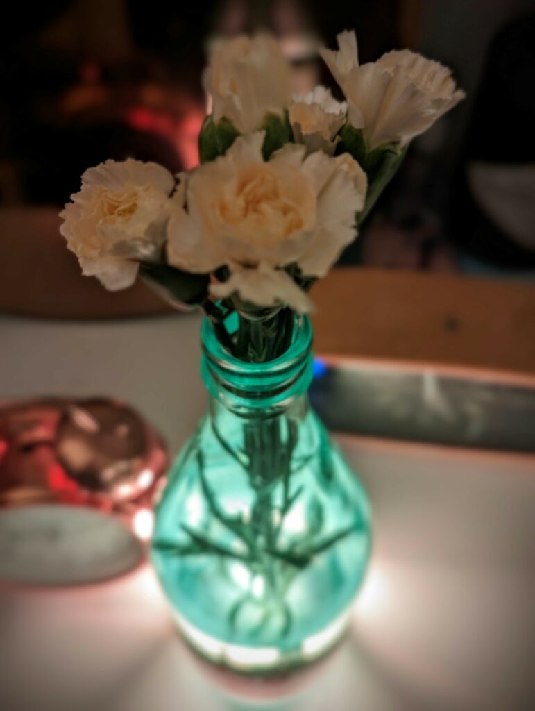 Creamy white carnations in a glowing turquoise glass vase, lit from below on a table with a warm, blurred background.