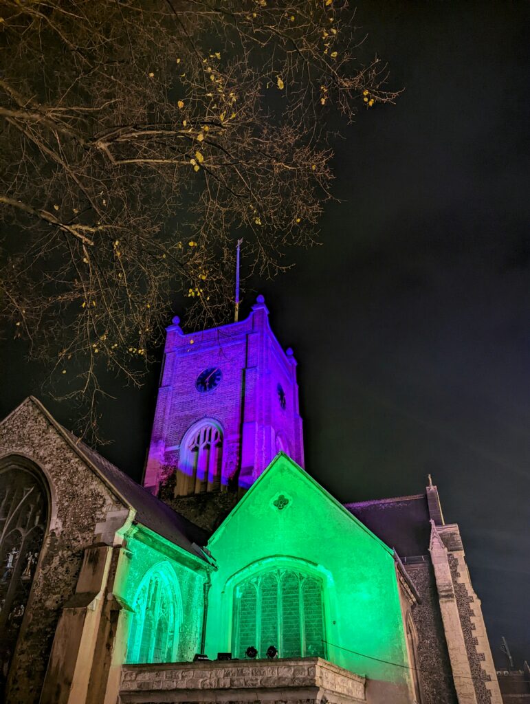 A stone church tower lit in vibrant purple and a lower facade in bright green at night, framed by bare branches against a dark sky.