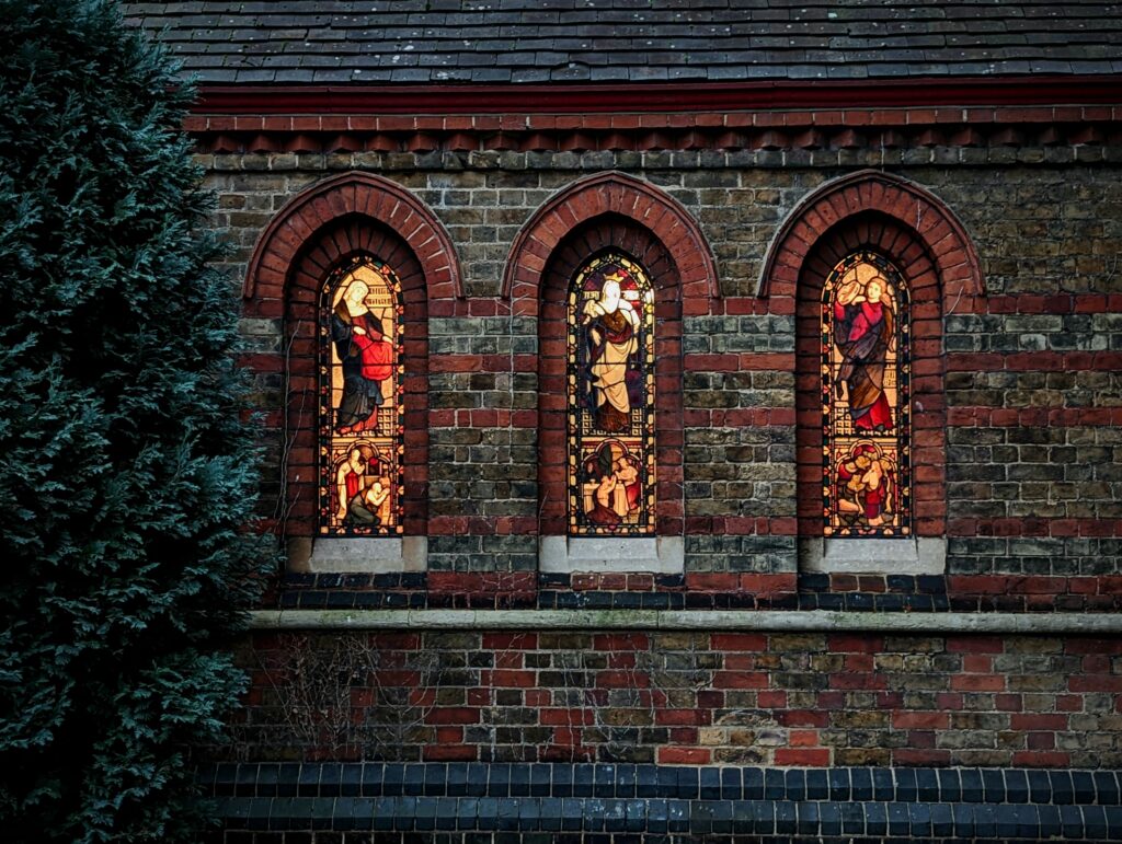 Three arched stained glass windows glowing from within a dark brick wall, next to a tall evergreen tree at night.