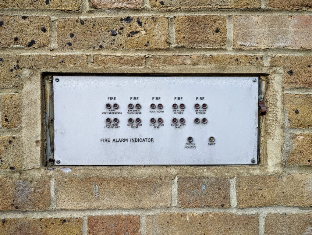 A metal fire alarm indicator panel with labeled lights for different building zones, recessed into a tan brick wall.