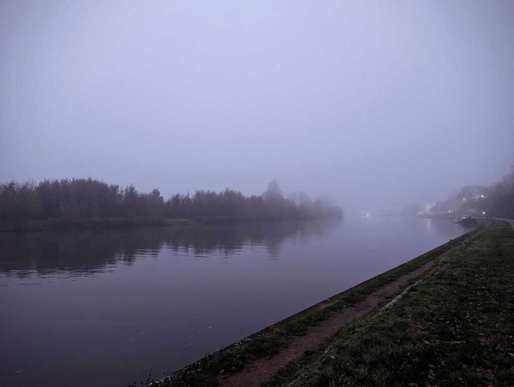 Thick fog obscuring a calm river at dusk, with a grassy path along the bank and dark trees reflected in the still water.