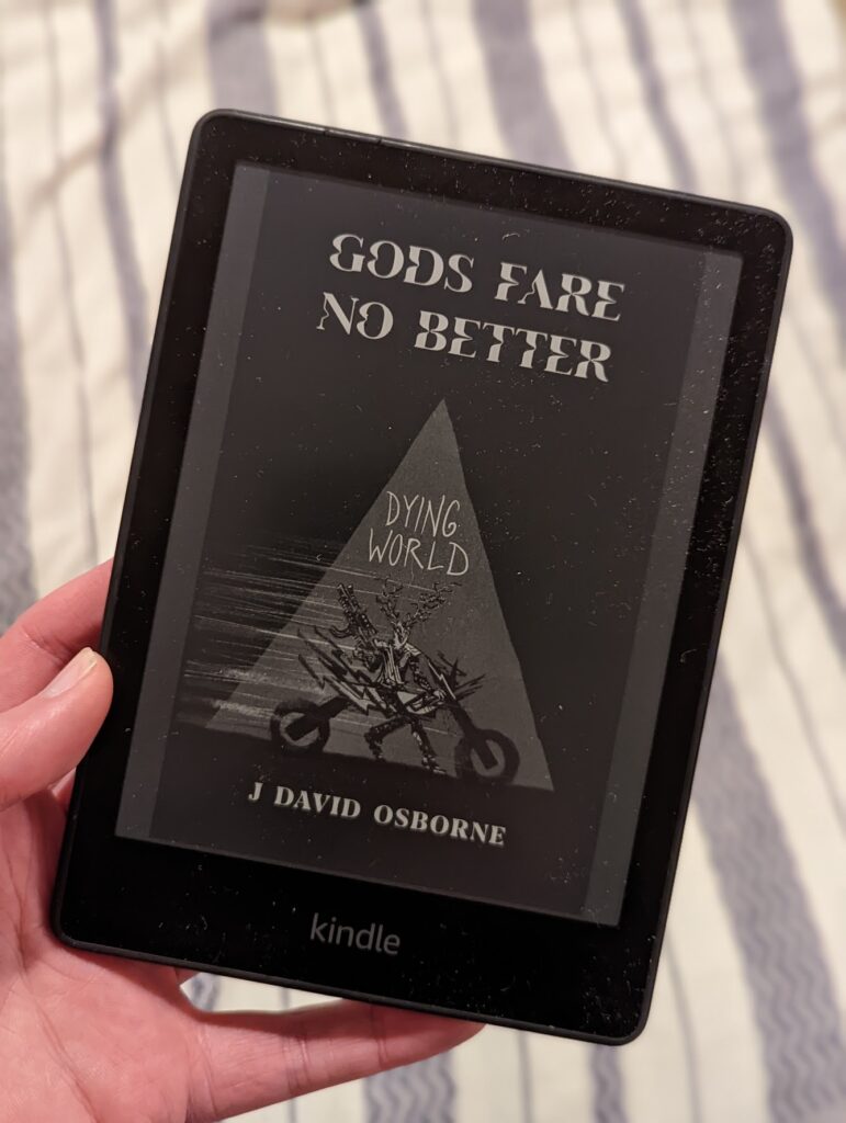 A hand holding a Kindle displaying the book cover for Gods Fare No Better by J David Osborne against a striped fabric background.