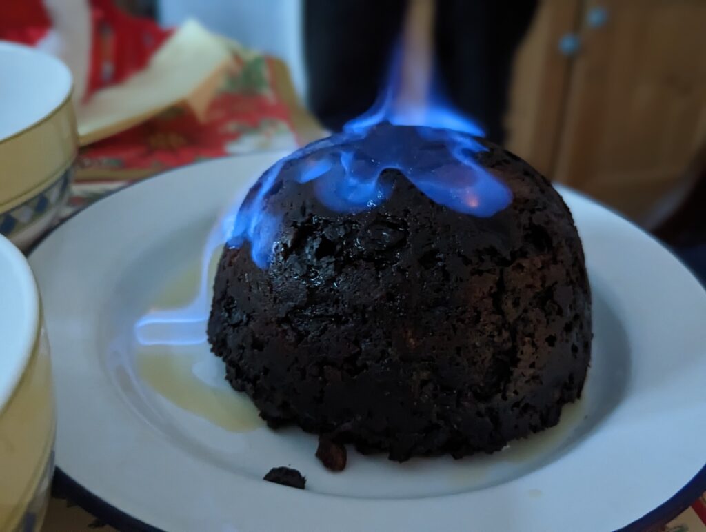 A round, dark Christmas pudding on a white plate with bright blue flames dancing across the top.