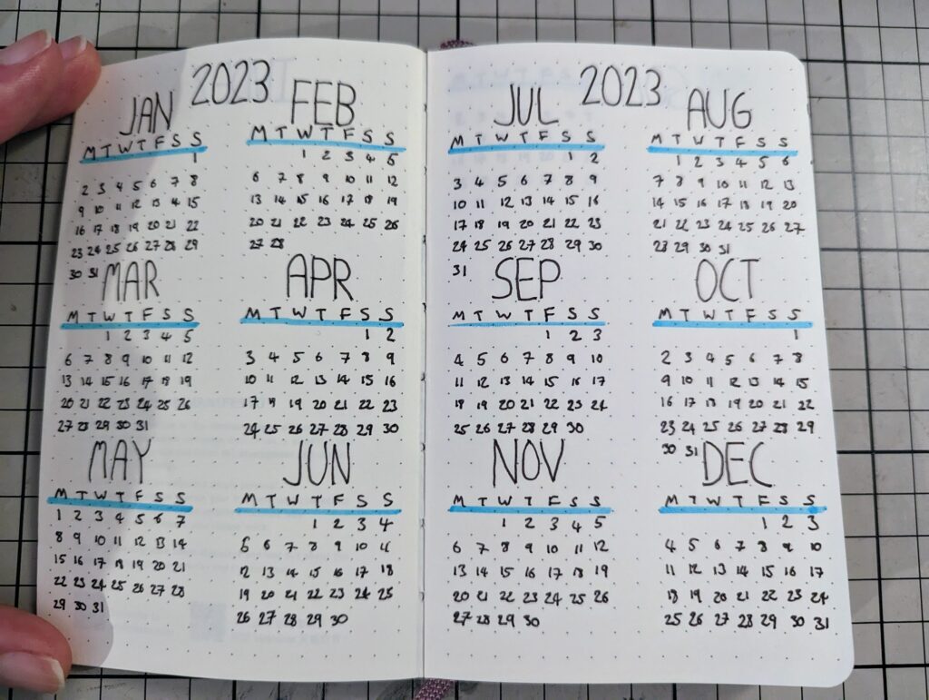 A hand-drawn 2023 calendar spread in a dot grid notebook, with blue highlighted headers and neatly written dates for all twelve months.
