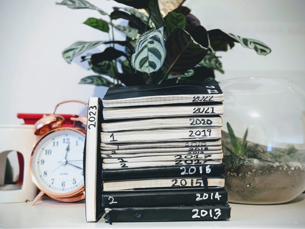 A stack of black journals with years 2013-2022 handwritten on the edges, a 2023 notebook standing upright beside a copper alarm clock and a plant.
