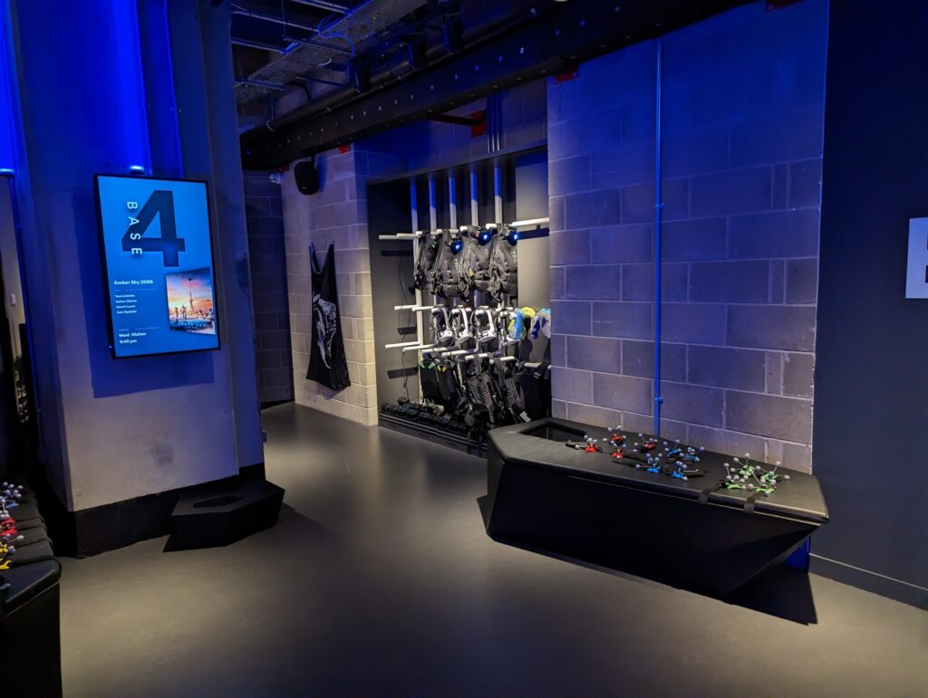 A blue-lit VR staging room with tactical vests on a rack, a Base 4 digital display, and motion capture markers on a black geometric bench.