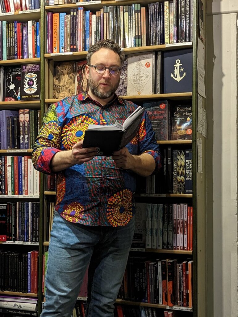 Brian Wilkins in a vibrant patterned shirt reading a book in front of packed bookshelves of magic and occult titles.