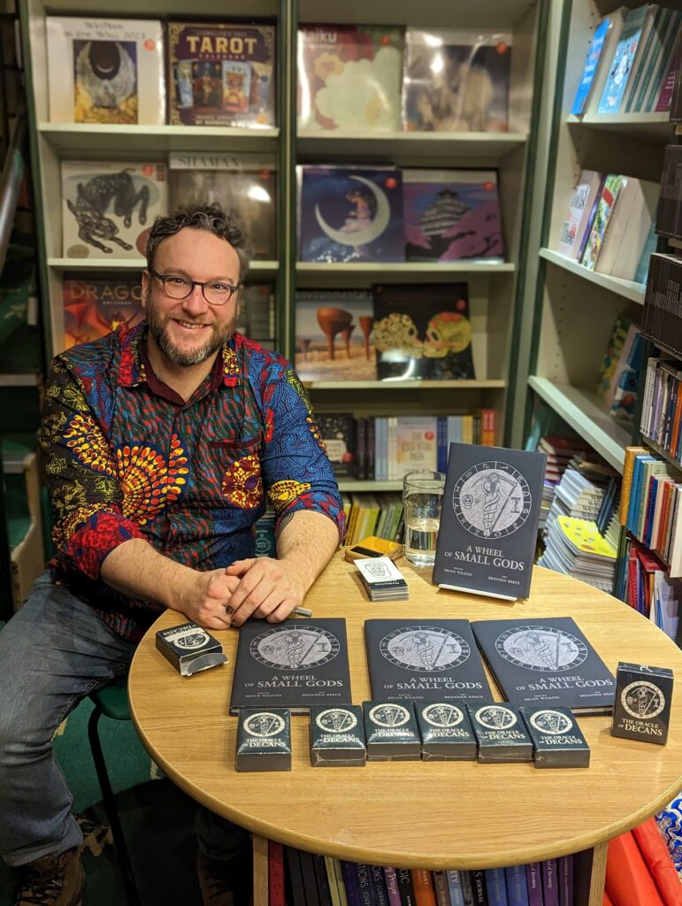 Brian Wilkins in a patterned shirt sits at a bookstore table displaying copies of A Wheel of Small Gods and The Oracle of Decans cards.