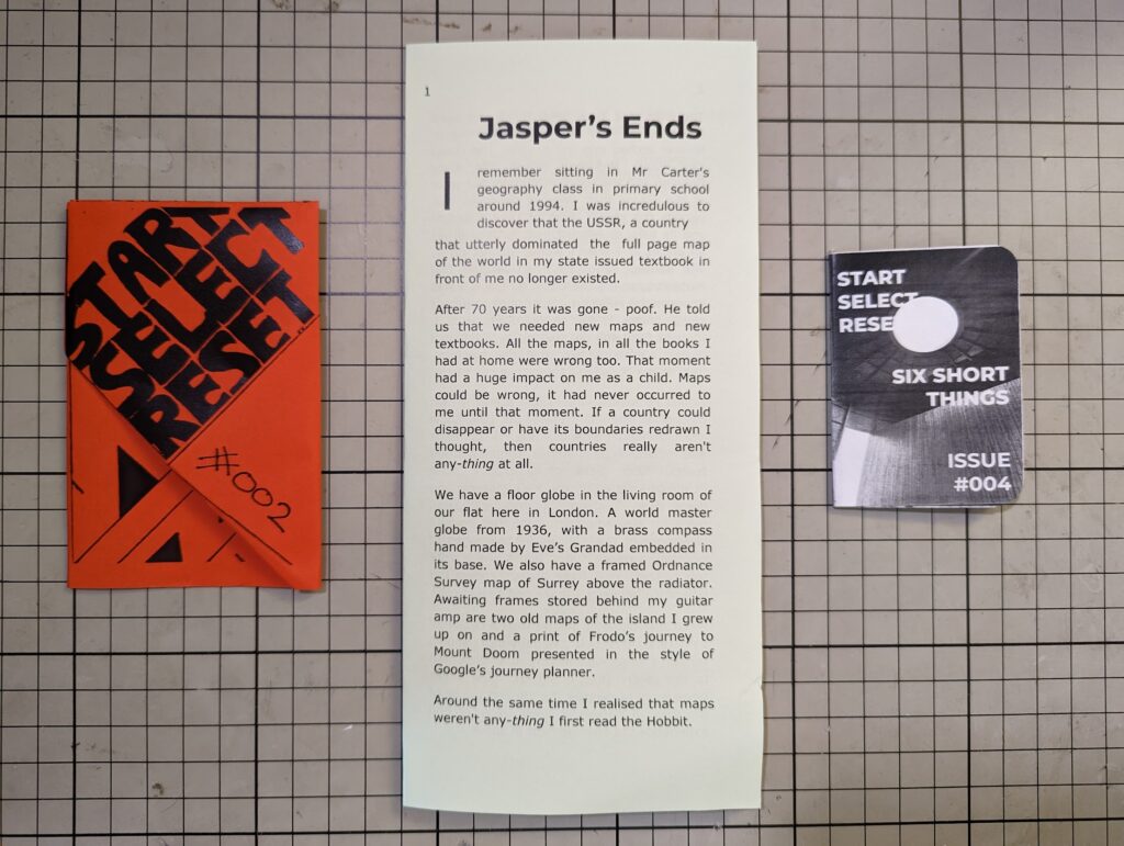 Featured image for Start Select Reset Zines 2022 - an orange zine, a text sheet, and a small booklet on a grid cutting mat.