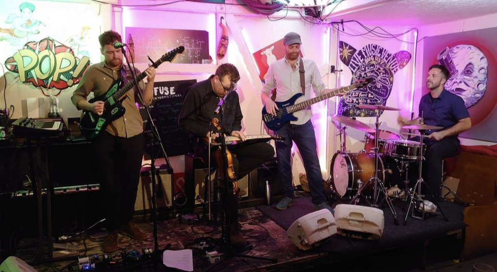 Four men performing live on a small stage with pink lighting, featuring a guitarist, lap steel player, bassist, and drummer.