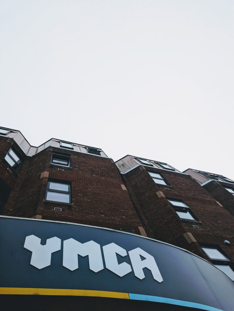 Low-angle view of a brick building with a white YMCA logo on a curved blue and yellow sign against a pale sky.